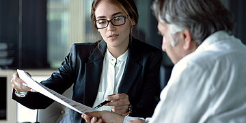 Eine Frau mit Brille und Jackett h&auml;lt Dokumente in den H&auml;nden, die sie ihrem Gegen&uuml;ber, einem &auml;lteren Mann, erkl&auml;rt.