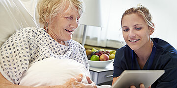 Foto: Eine &auml;ltere Frau liegt im Bett und schaut mit einer j&uuml;ngeren Frau auf ein Tablet.