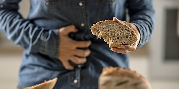Zöliakie Im Vordergrund liegt ein Brot. Im Hintergrund ein Mann, der sich seinen schmerzenden Bauch hält, in der Hand eine Scheibe des Brotes.