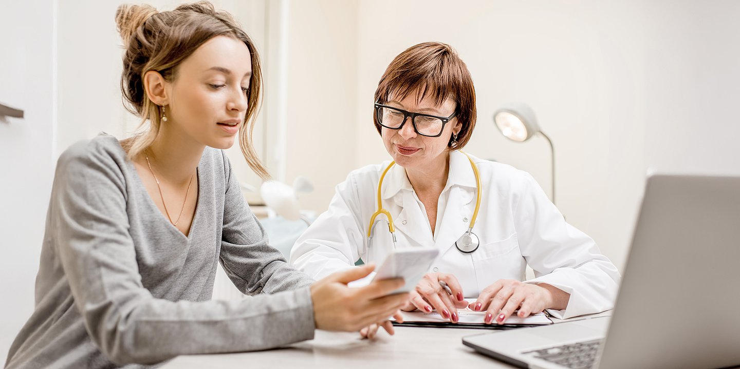 Foto: Eine Ärztin und ihre Patientin sitzen im Behandlungsraum und schauen gemeinsam auf ein Handy.