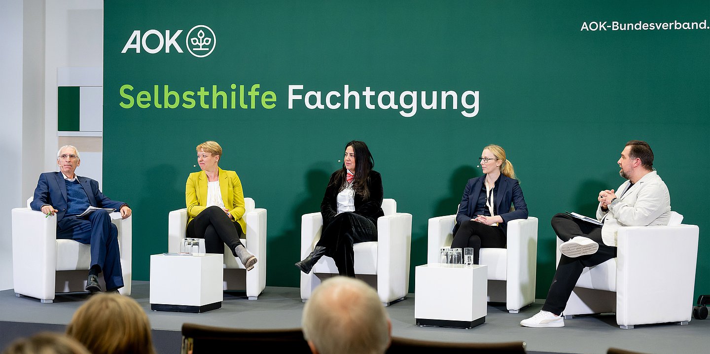 Auf einem Podium sitzen in weißen Ledersesseln fünf Männer und Frauen und sind in ein Gespräch vertieft.  