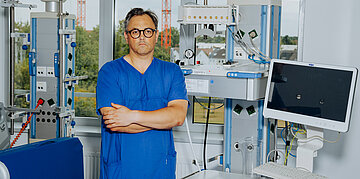 Foto: Der Arzt Christian Karagiannidis steht in blauer Klinikkleidung zwischen medizinischen Ger&auml;ten einer Intensivstation.