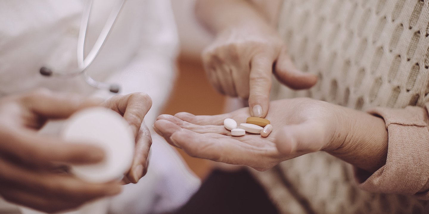 Foto: Eine ältere Person hält Tabletten in der Hand und zeigt auf eine davon. Eine zweite Person mit Stethoskop hat eine Tablettendose in der Hand.