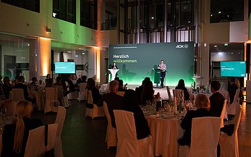 Foto: Ein Saal mit Zuschauern. Sie blicken in Richtung Bühne, auf der zwei Personen eine Rede halten.