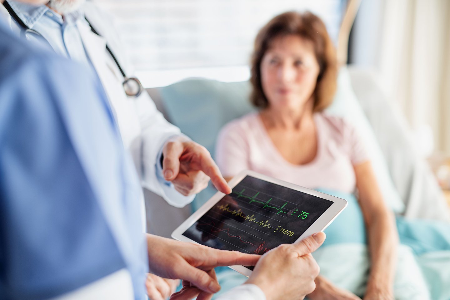 Das Foto zeigt im Hintergrund eine Frau, die im Krankenhaus im Bett sitzt und zum Arzt schaut, der im Vordergrund mit einer anderen Person auf ein Tablet schaut und mit dem Finger auf einen bestimmten Wert zeigt.