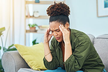 Eine junge Frau sitzt leicht vorgebeugt auf einem Sofa. Die Augen sind geschlossen, das Gesicht wirkt angespannt und sie fasst sich mit beiden H&auml;nden an den Schl&auml;fen. Die Arme sind dabei auf den Knien abgest&uuml;tzt.