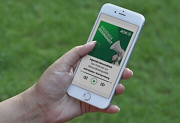 Eine Hand h&auml;lt ein Smartphone, auf dessen Display der neue Podcast zu sehen ist.
