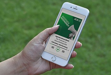 Eine Hand hält ein Smartphone, auf dessen Display der neue Podcast zu sehen ist.