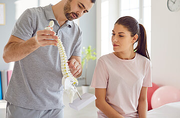 Eine junge Frau sitzt beim Physiotherapeuten im Behandlungsraum, der neben ihr steht und ihr an einem Wirbels&auml;ulenmodell etwas zeigt und erkl&auml;rt.