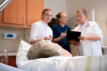 Ein &auml;lterer Patient liegt im Krankenhausbett, w&auml;hrend zwei &Auml;rztinnen und ein Arzt im Hintergrund eine Besprechung mit einem Tablet f&uuml;hren.