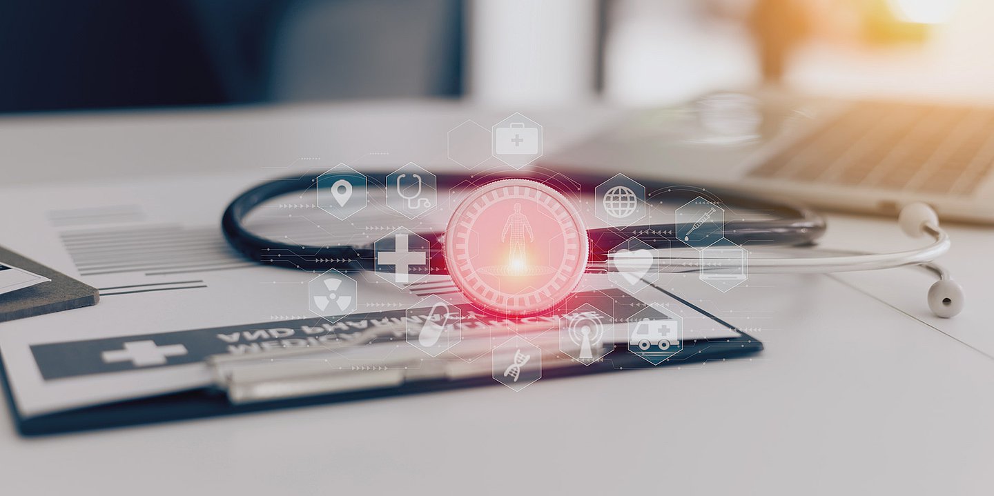 Foto: Ein Stethoskop liegt neben einem Laptop und einem Klemmbrett und leuchtet – um das Stethoskop sind verschiedene Gesundheitssymbole gruppiert.