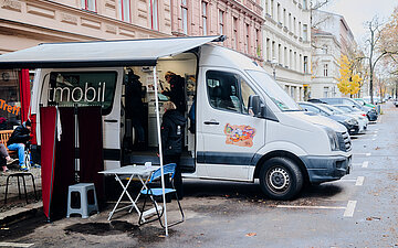 Das Arztmobil an seinem Standort: Ein Van, die Seitent&uuml;r offen, daneben eine &Uuml;berdachung mit Tisch, St&uuml;hlen und einem Sichtschutz.