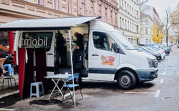 Das Arztmobil an seinem Standort: Ein Van, die Seitentür offen, daneben eine Überdachung mit Tisch, Stühlen und einem Sichtschutz.