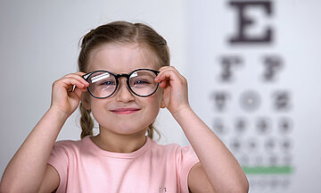 Foto zeigt ein M&auml;dchen im Grundschulalter beim Optiker, wo es eine Brille aufsetzt und dabei l&auml;chelt. Im Hintergrund sind die an der Wand projezierten Buchstaben eines Sehtest zu sehen.