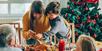 Weihnachten Eine Familie beim Festtagsessen, eine junge Frau und ein Kind verteilen lachend Essen, ein geschmückter Weihachtsbaum steht im Hintergrund.