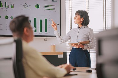 Foto: Eine Frau erkl&auml;rt Grafiken an einem Whiteboard. Im Vordergrund sitzt ein Mann.