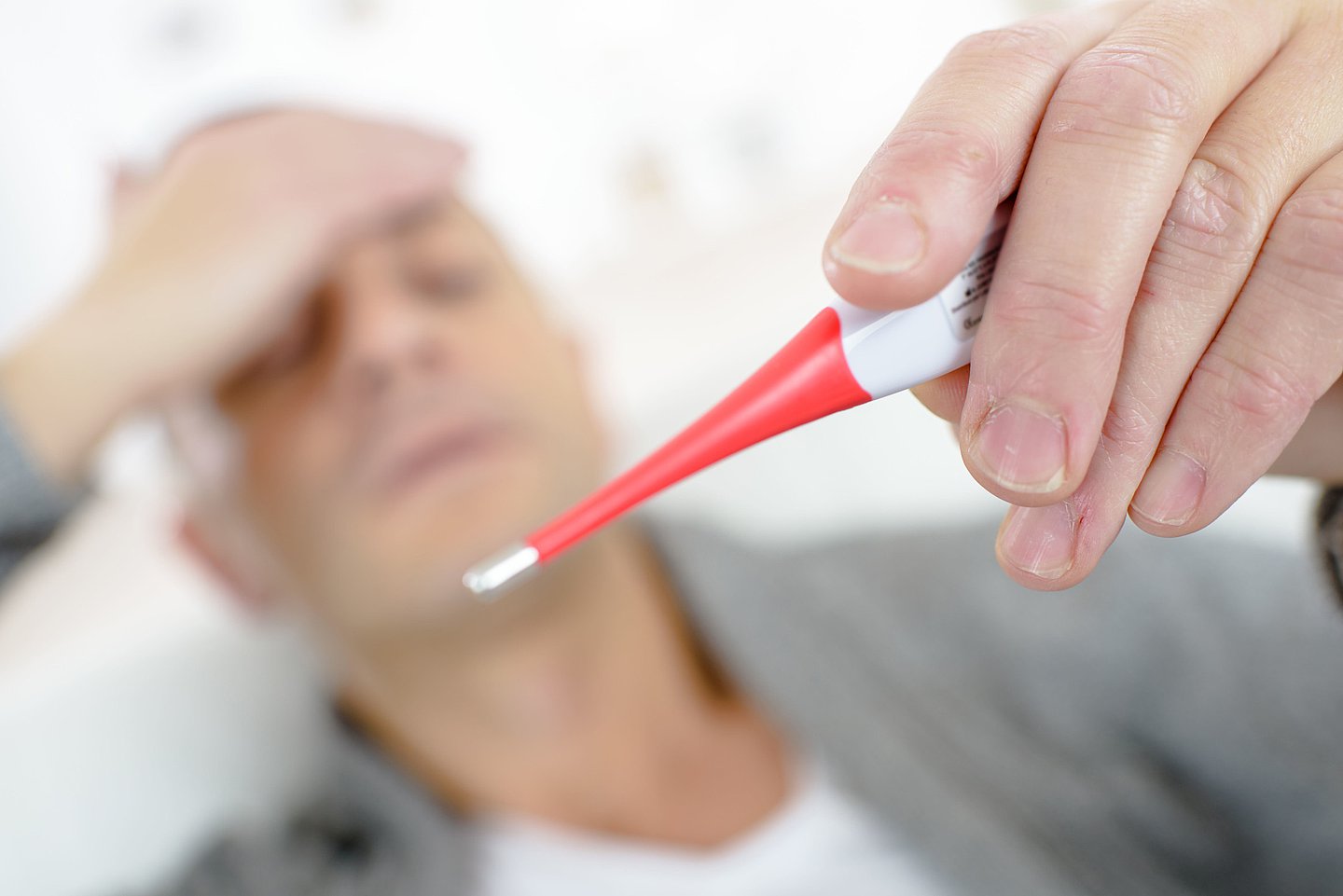 Ein liegender Mann fasst sich an die Stirn und schaut auf ein Fieberthermometer.