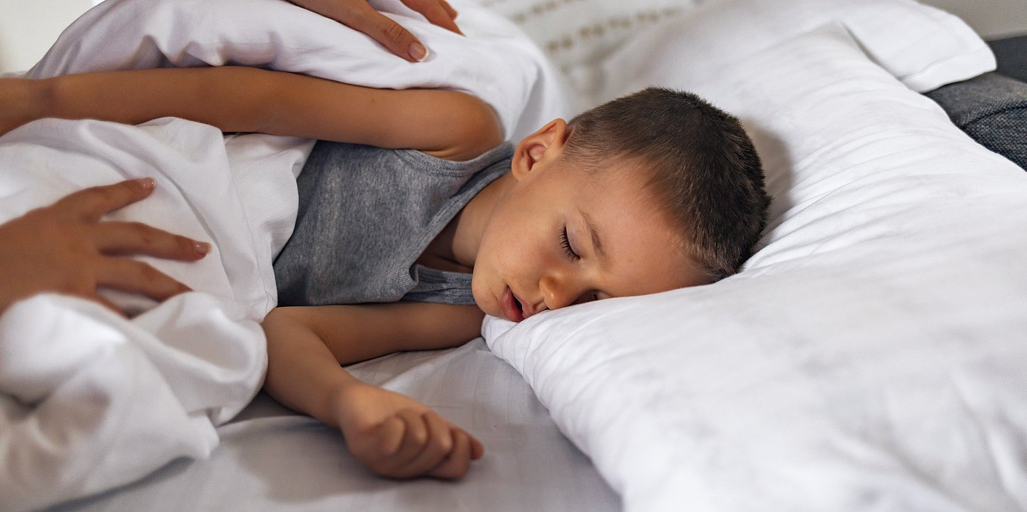 Ein kleiner Junge liegt schlafend im Bett, Eltern wecken ihn. 