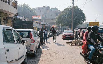 Eine Straße in Indien. Rechts und links parken Autos, weiter hinten sind Läden. Auf der Straße stehen verschiedene Personen. Vorne rechts fahren zwei Menschen auf einem Motorrad.