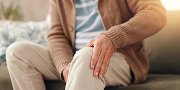DMP Osteoporose und Rheuma Auf einer Couch sitzt eine Person und hält sich das schmerzende Knie.