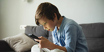 ein kleiner Junge sitzt auf einem Sofa und schaut auf ein Smartphone.