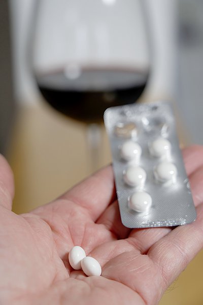 Eine Person h&auml;lt ihrer Hand Medikamente. Im Hintergrund steht ein Glas Wein auf dem Tisch.