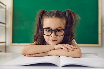 Das Foto zeigt ein Mädchen in der Schulklasse im Nahformat, das eine Brille trägt, vor einer Tafel an ihrem Tisch mit Heft.
