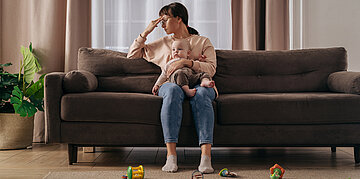 Eine Frau sitzt mit einem Baby auf der Couch, blickt ersch&ouml;pft und fasst sich an den Kopf.