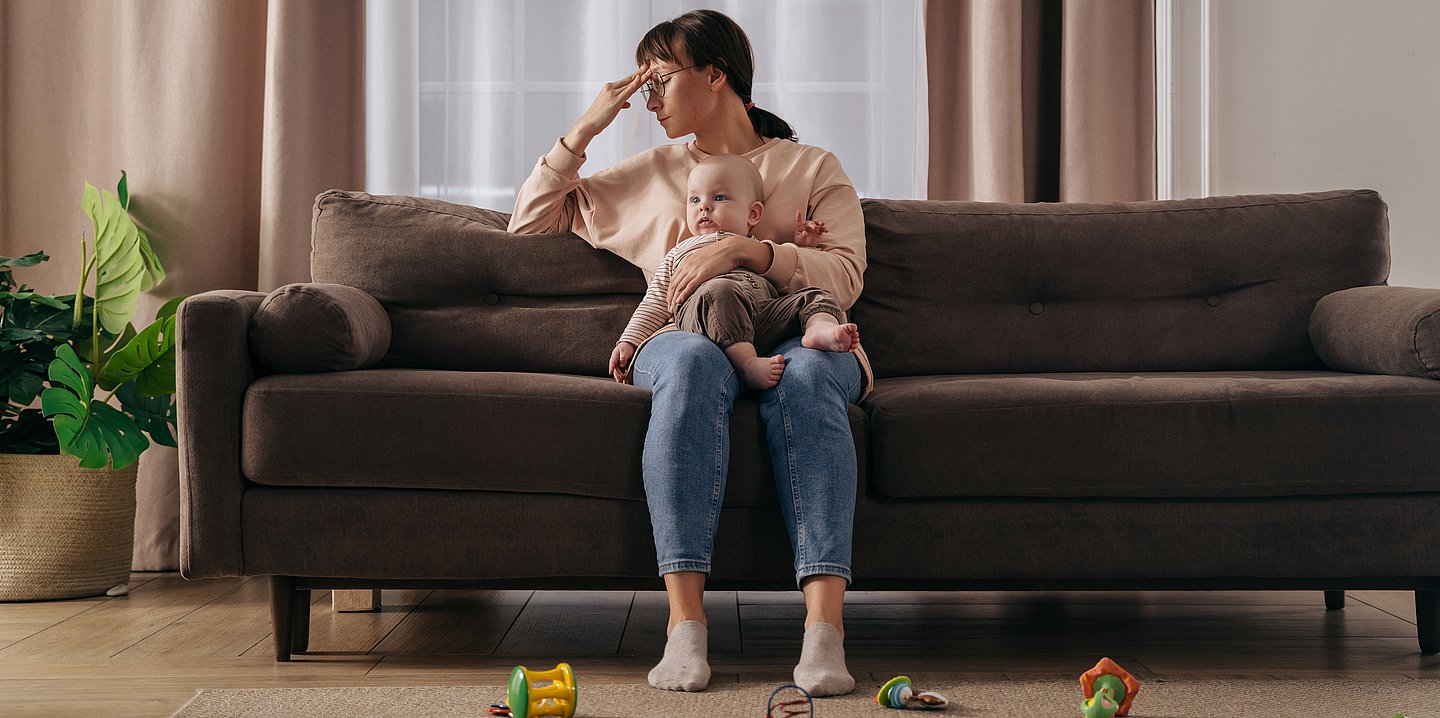 Eine Frau sitzt mit einem Baby auf der Couch, blickt erschöpft und fasst sich an den Kopf.