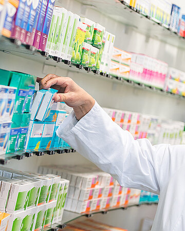 Eine Person in einem wei&szlig;en Kittel greift in einem Apothekenregal nach einer Medikamentenpackung.