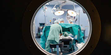 Foto: Durch ein Bullauge ist ein OP-Saal zu sehen, in dem &Auml;rzte und &Auml;rztinnen eine Patientin oder einen Patienten operieren.
