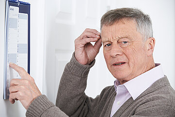 Ein &auml;lterer Mann steht vor einem Kalenderblatt, das an der Wand befestigt ist. Die dem Zeigefinger der linken Hand zeigt er auf einen speziellen Tag im Kalender, mit der rechten ber&uuml;hrt er seine rechte Schl&auml;fe und schaut etwas ratlos.