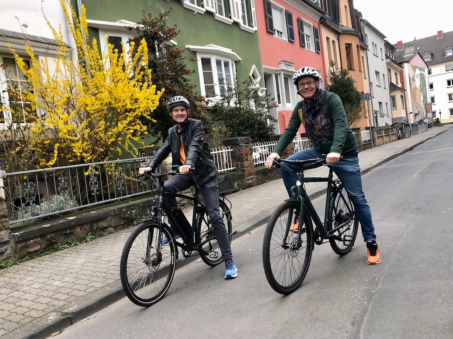 Foto: Zwei Männer nebeneinander auf jeweils auf einem E-Bike sitzend vor einer Srraßenkulisse mit bunten Häuschen, Vorstadtatmosphäre