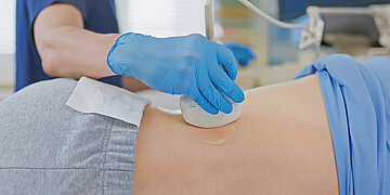 Foto: Medizinische Untersuchung mit Ultraschallsonde am seitlichen Bauch einer liegenden Person; eine medizinische Fachkraft tr&auml;gt blaue Handschuhe.
