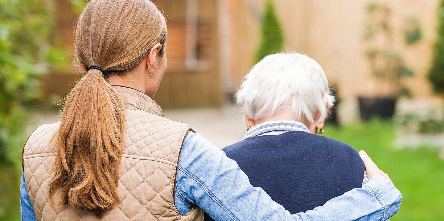 Foto: Eine ältere und eine jüngere Frau von hinten zu sehen, laufen nebeneinander. Die jüngere Frau umarmt die ältere.