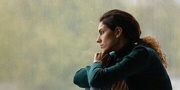 Eine Frau sitzt zusammengekauert vor einer gro&szlig;en Fensterscheibe. Drau&szlig;en regnet es. Sie starrt mit ausdruckslosem Gesicht ins Nichts.