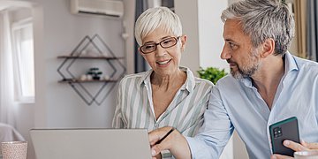 Paar vor dem Laptop Das Bild zeigt einen Mann (rechts) und eine Frau am Küchenetisch vor einem Laptop bei der Recherche. Die Frau hat nahezu weiße Haare und trägt eine hellgrau-weiß-länsgestreifte Bluse. Der Mann hat graumeliertes Harr und einen Vollnart. Sein Hemd ist hellblau, fast weiß. In seiner linken Hand hält er ein Smartphone.