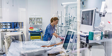 In einem Krankenhauszimmer mit vielen verschiedenen technischen Ger&auml;ten pr&uuml;ft eine Krankenpflegerin die Elektroden an einem Patienten oder einer Patientin.