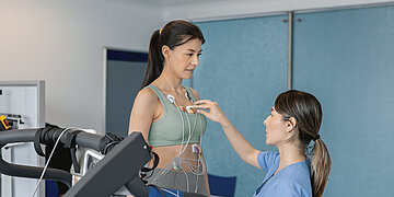 Eine Frau steht auf einem Laufband und ist mit mehreren Elektroden am Oberk&ouml;rper verkabelt, w&auml;hrend eine medizinische Fachkraft in blauer Kleidung die Sensoren &uuml;berpr&uuml;ft.