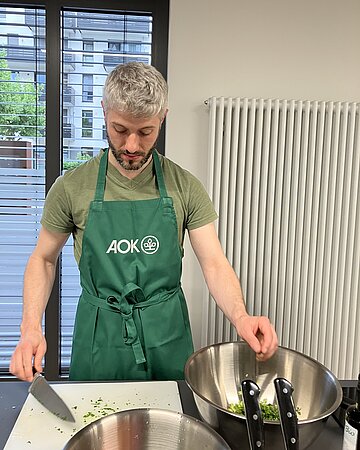 Ein Mann mit AOK-Sch&uuml;rze steht vor zwei gro&szlig;en Sch&uuml;sseln und bereitet Essen zu.