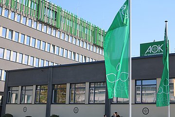 AOK-Zentrale in Bad Homburg Das Bild zeigt ein großes grau-braunes Verwaltungsgebäude im Anschnitt mit drei parallelen Fensterreihen mit. Oben schließt der Bau mit grünen Längsstreben ab. Vor dem Hauptgebäude steht eine Art Büro-Pavillion, auf dem Dach ein AOK-Schild, davor zwei Masten mit grünen AOK-Fahnen.