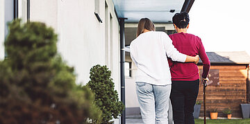Eine j&uuml;ngere Person unterst&uuml;tzt eine &auml;ltere Person mit Gehstock beim Gehen auf einem Weg vor einem Wohnhaus.