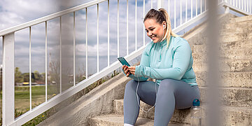 Foto einer &uuml;bergewichtigen Frau im Sportoutfit, die l&auml;chelnd auf einer Au&szlig;entreppe sitzt und in ihr Smartphone blickt.