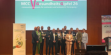 Auf einer B&uuml;hne stehen zw&ouml;f Leute, die Blumenstr&auml;u&szlig;e in den H&auml;nden halten.