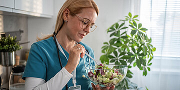 Eine blonde Frau mit Brille in Pflegekleidung steht in einer K&uuml;che und isst Salat.