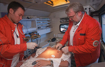 Das Bild zeigt zwei Rettungssanit&auml;ter mit orangen Rettungsjacken beim EKG im Rettungswagen. In der Mitte liegt ein Patient mit Sauerstoffmaske, dem die Pads des EKG angelegt werden. Am linken Finger tr&auml;gt er einen Pulsmesser.