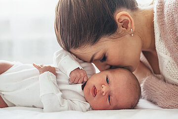 Das Foto zeigt ein Baby auf einer Unterlage, die Mutter beugt sich von der Kopfseite her &uuml;ber ihr Baby und liebkost es seitlich am Kopf.und gt