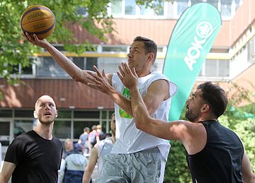 Vier Sportler im Vordergrund, einer davon im Sprung h&auml;lt einen Basketball in der rechten Handfl&auml;che. 