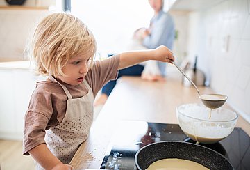 Verbrennungen bei Kindern in Westfalen-Lippe Ein kleines Kind steht am Herd und füllt mit einer Kelle Teig aus einer Schale in die Pfanne, die auf dem Kochfeld steht.