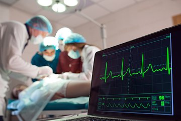 Ärzte operieren einen Patienten, im Vordergrund werden die Vitalwerte auf einem Monitorangezeigt.